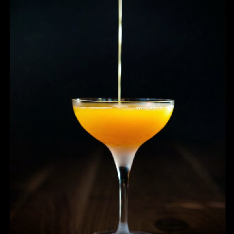 Golden liquid pouring into an orange cocktail in a frosted coupe glass.