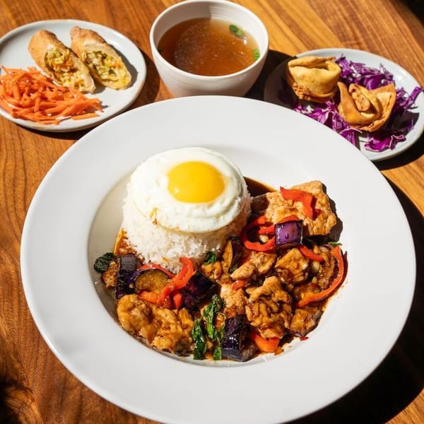 Thai basil chicken with fried egg, rice, soup, spring rolls, and fried wontons on a wooden table.