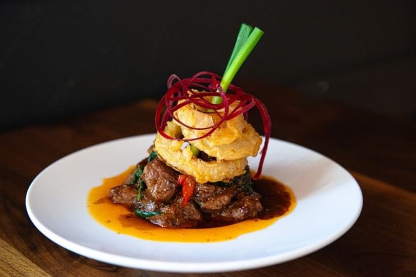 Braised meat dish with a rich sauce, topped with crispy onion rings, red garnish, and green onion.
