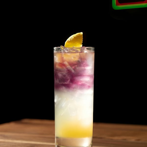 Layered cocktail with yellow, white, and purple liquids, garnished with a lemon slice on a wooden table.