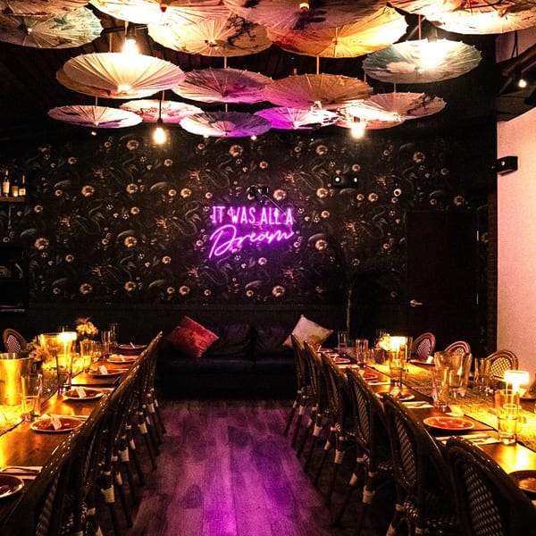 Atmospheric restaurant dining room with long tables, paper umbrellas, and a glowing "It Was All A Dream" sign.