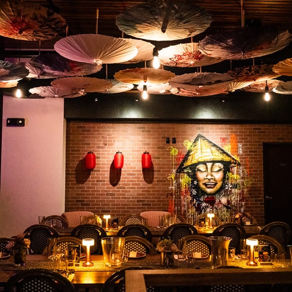 Asian-themed restaurant interior with ceiling umbrellas, set tables, and a mural of a person in a conical hat.