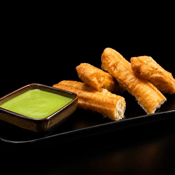 Golden fried dough sticks with a bowl of green dipping sauce on a black plate.