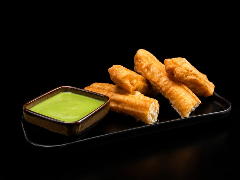 Golden fried dough sticks with a bowl of green dipping sauce on a black plate.